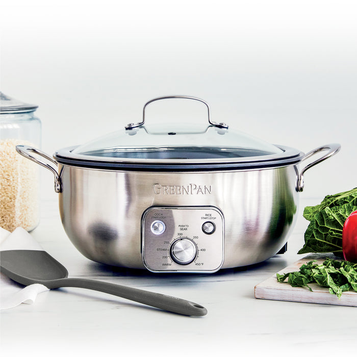 Electric wok with black exterior on a kitchen counter, next to assorted vegetables, grains, and a spatula.