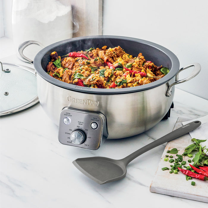 Electric wok with meat and vegetables, a spatula inside, on a white countertop.