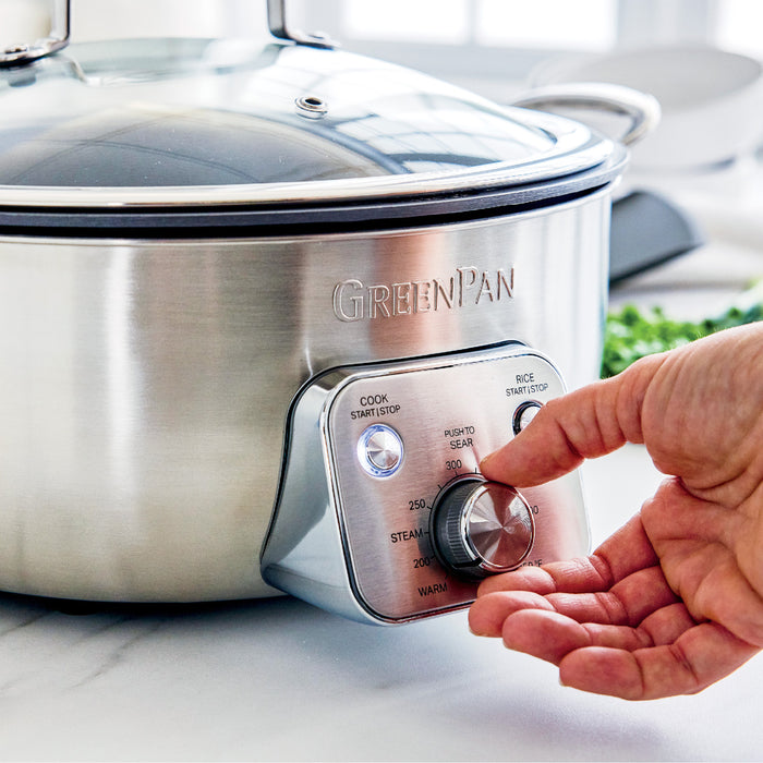Hand adjusting temperature dial on stainless steel electric wok with black handles, 6-quart size visible.