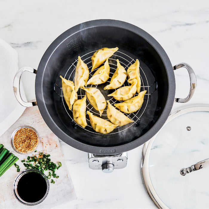 Electric wok steaming dumplings on a rack with green onions, soy sauce, and sesame seeds arranged beside it.