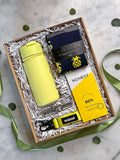A curated gift box set on a marble surface, featuring a bright yellow insulated bottle, navy socks with yellow pineapples, a yellow Honest brand soap bar, and a yellow and black Sealand keychain, all arranged in white paper shred inside a wooden box.