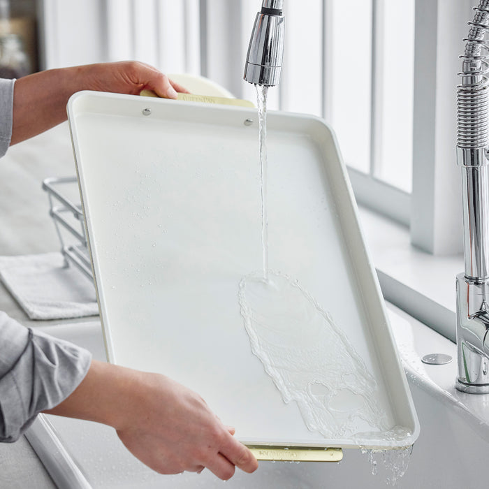 A 13 x 18 metal half sheet pan with cooling rack being rinsed under running water in a modern kitchen sink.