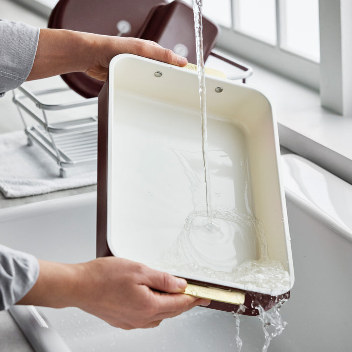 Reserve Bakeware 13″ x 9″ rectangular pan in Bordeaux with a cream interior being rinsed under a kitchen faucet.