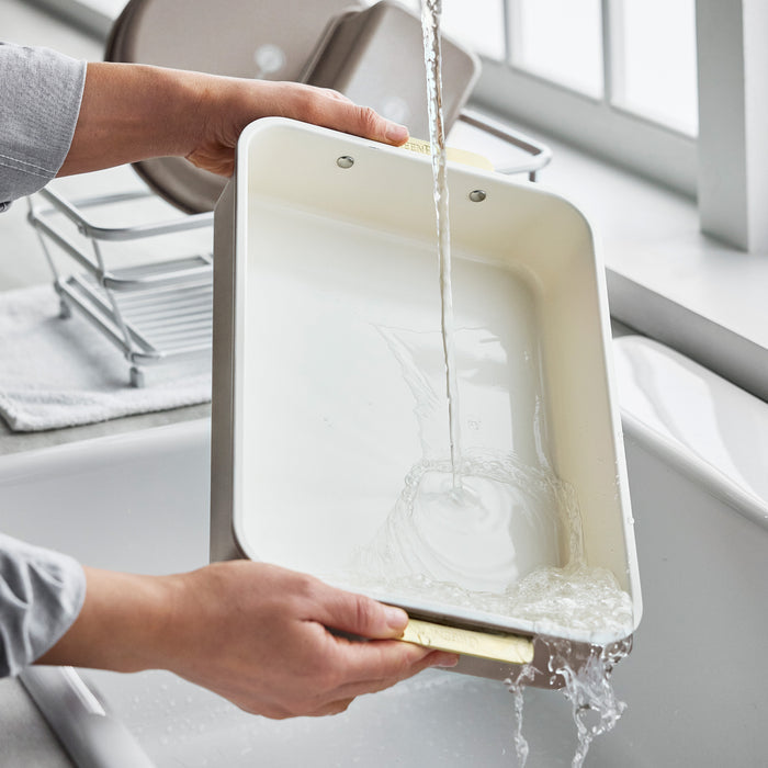 Hands rinsing a latte-colored Reserve Bakeware 13″ x 9″ rectangular pan under a faucet.