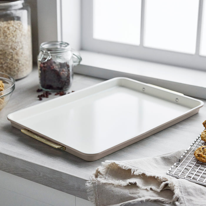 Reserve Bakeware 13″ x 18″ half sheet pan in latte, featuring light beige aluminized steel and gold-toned handles on a counter.