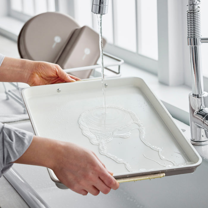 Hands rinsing a 13″ x 18″ Latte Reserve Half Sheet Pan under a kitchen faucet.