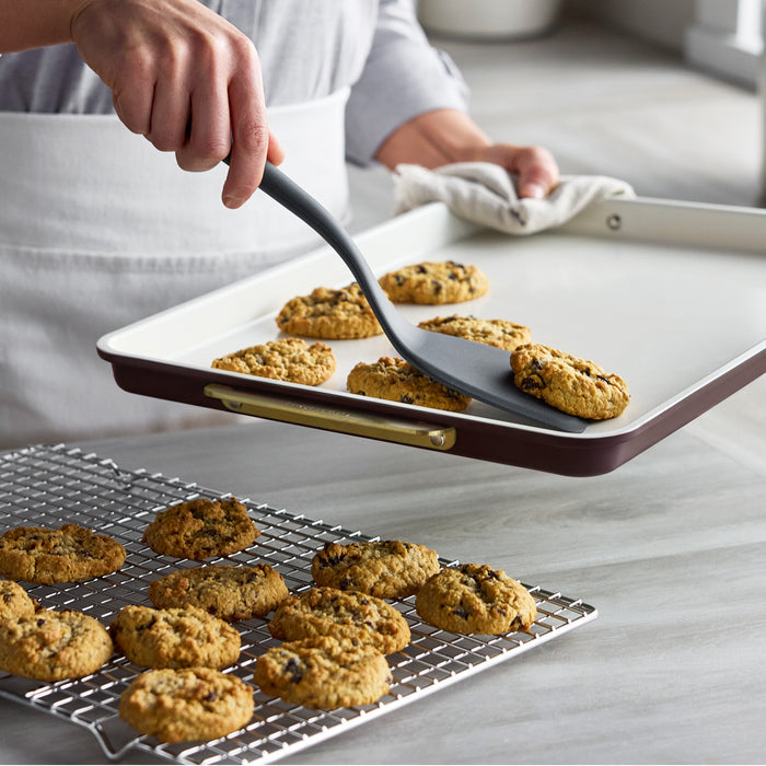 Spatula moving an oatmeal cookie from a Bordeaux Reserve Bakeware 7-piece set baking sheet to a cooling rack.