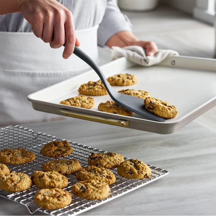 Cookie being moved from a Latte Reserve Bakeware 7-piece aluminized steel baking sheet to a cooling rack with a grey spatula.