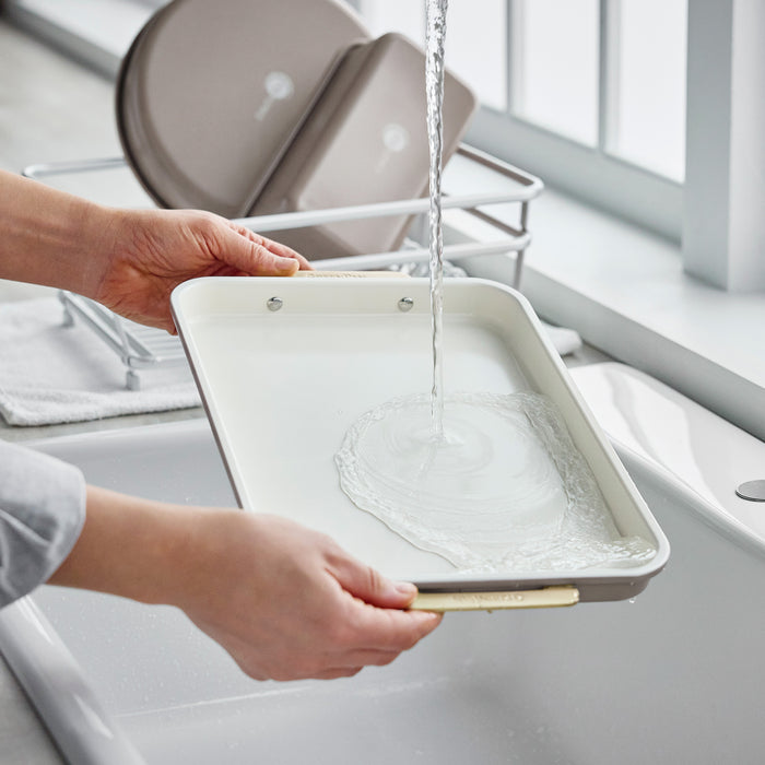 Hands rinsing a latte-colored, 8-piece aluminized steel Reserve Bakeware set under a kitchen faucet.