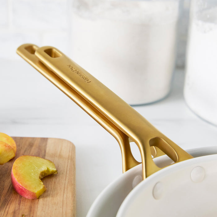 Cream frypans with gold handles on a white surface with apples and a cutting board.
