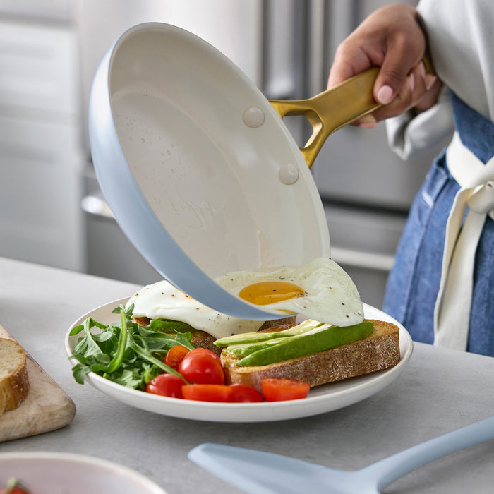 Person pouring food from a light blue frying pan onto a plate with a sandwich and salad.