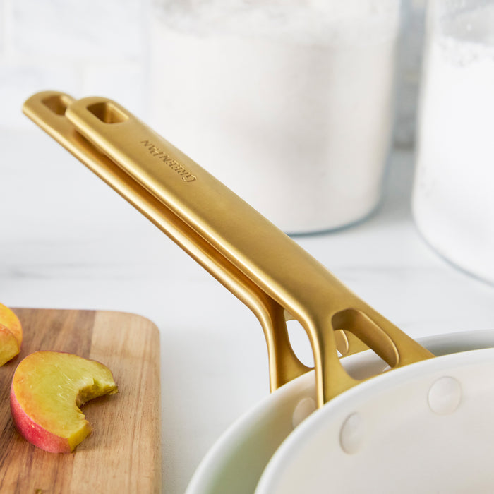 Taupe frypans with gold handles on a white surface with apples and a cutting board.