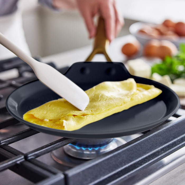 Hand folding an omelet in a black ceramic nonstick pan from a 14-piece set on a gas stove.