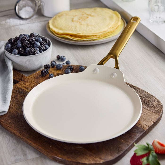 Crepe pan with gold handle on a wooden cutting board, surrounded by crepes and berries.