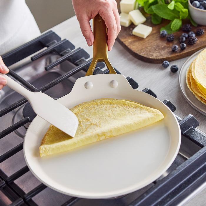 Folding a crepe in a ceramic nonstick pan; butter, berries, and mint are arranged on a board beside the stove.