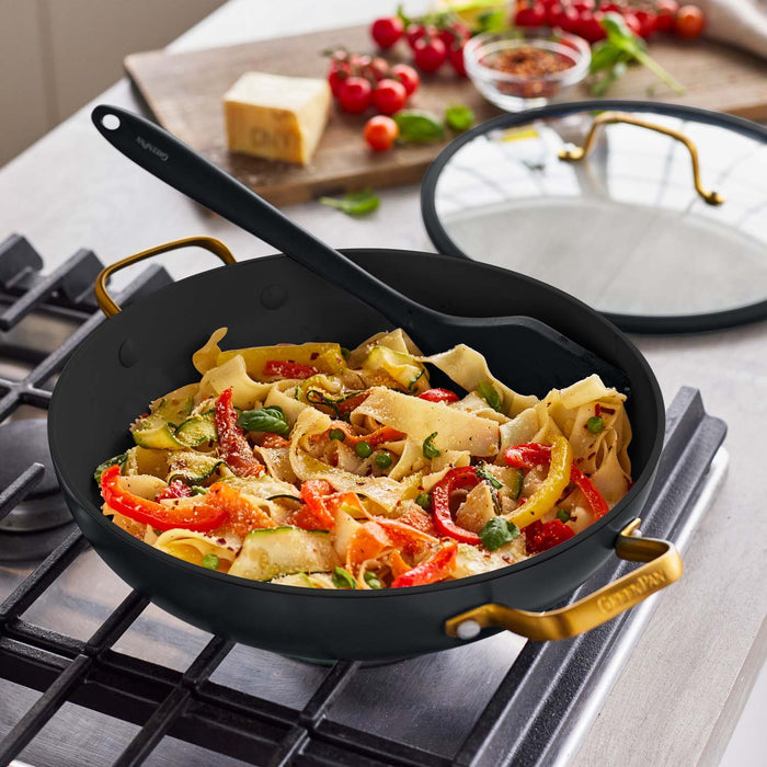 Black ceramic nonstick pan with lid and spatula on stove, cooking pasta with vegetables and herbs.