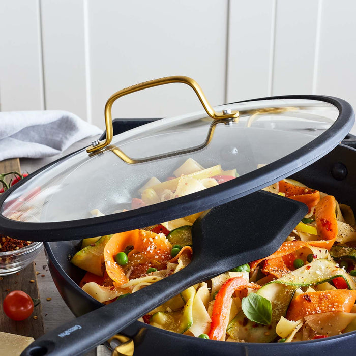 Black ceramic nonstick skillet with pasta, vegetables, glass lid, and spatula resting on the pan.