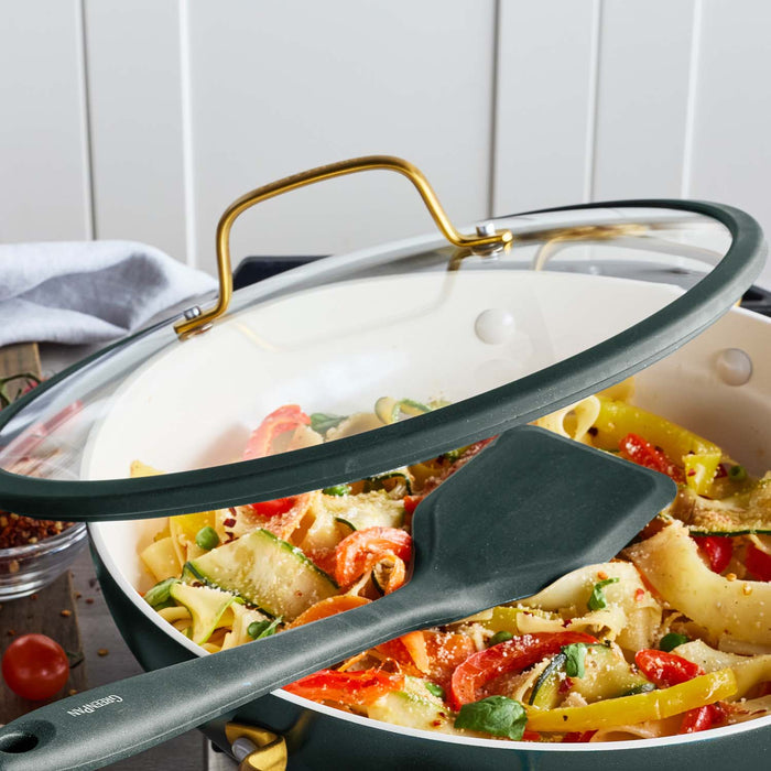 Ceramic spatula rests on a crepe pan from a 14-piece cookware set, with pasta, vegetables, herbs, and cheese inside the pan.