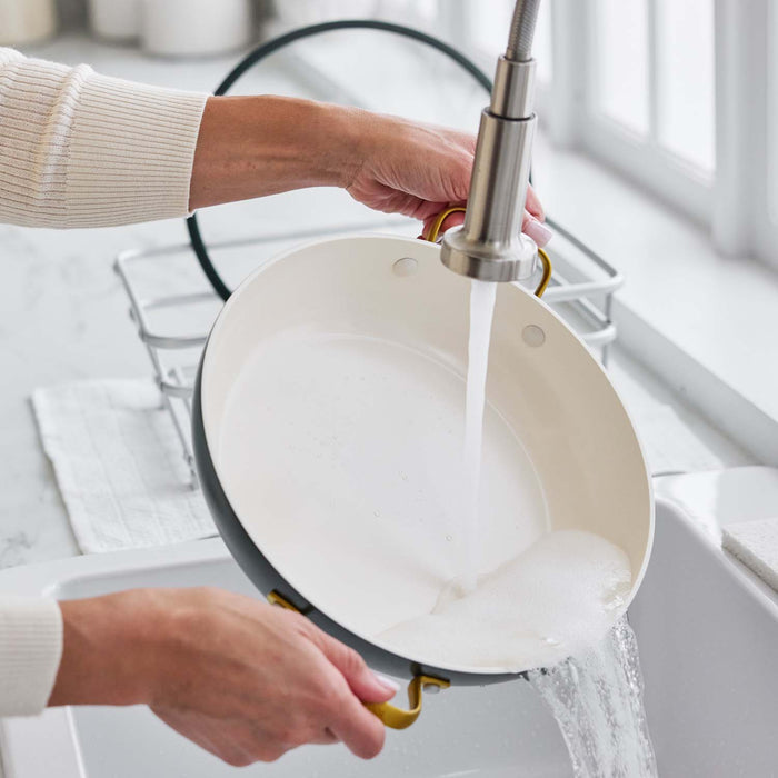 Washing a green ceramic nonstick pan with lid under running water by a sudsy sink and drying rack.