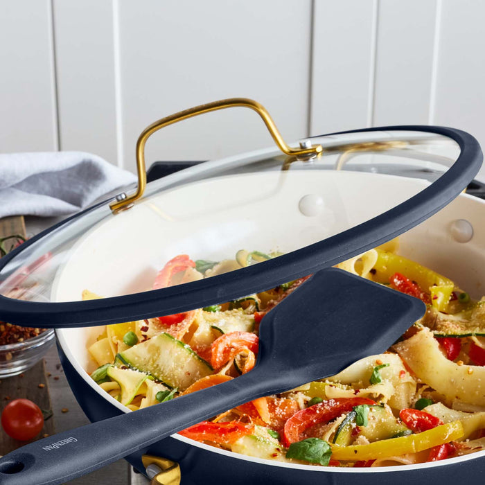 Blue ceramic nonstick cookware set with pasta in a skillet, spatula, and clear lid on a wooden surface.