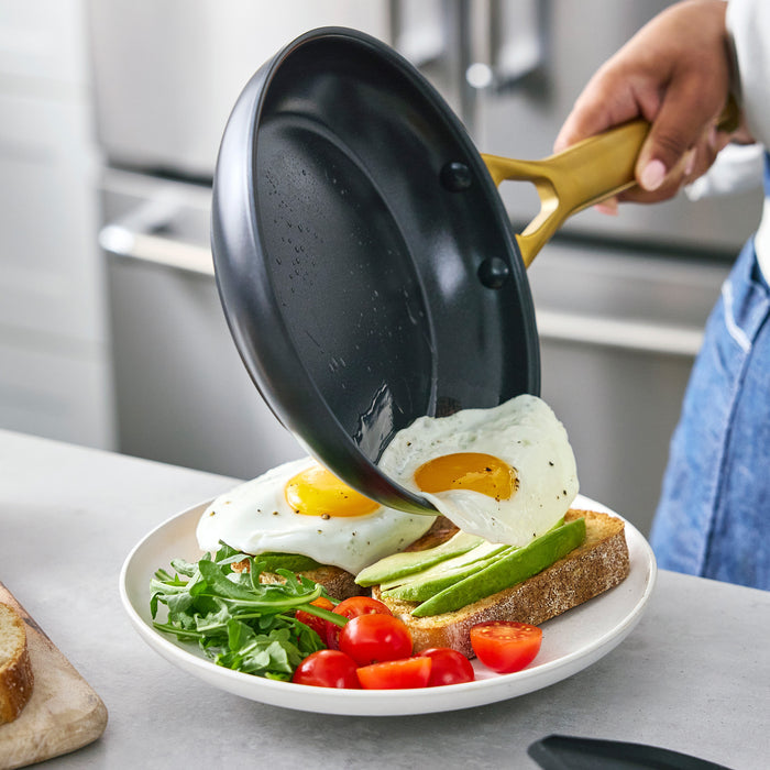 Person pouring eggs from a black frying pan onto a plate with toast, avocado, and tomatoes in a kitchen setting.