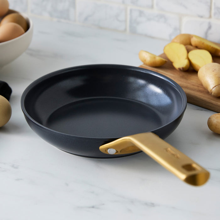 Black frying pan with a gold handle on a kitchen counter with ingredients in the background.