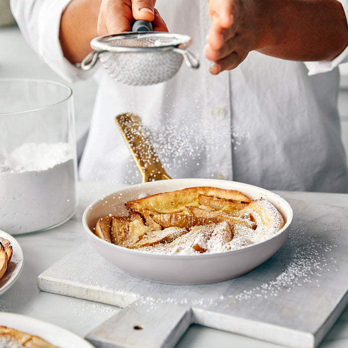 Person dusting powdered sugar over a dish of baked apple slices on a marble surface.