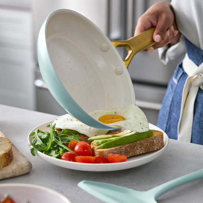Person holding a light blue frying pan with a fried egg over a plate of toast and salad.