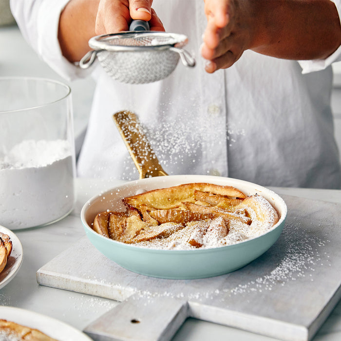 Person dusting a dish of baked apple dessert with powdered sugar on a kitchen counter.