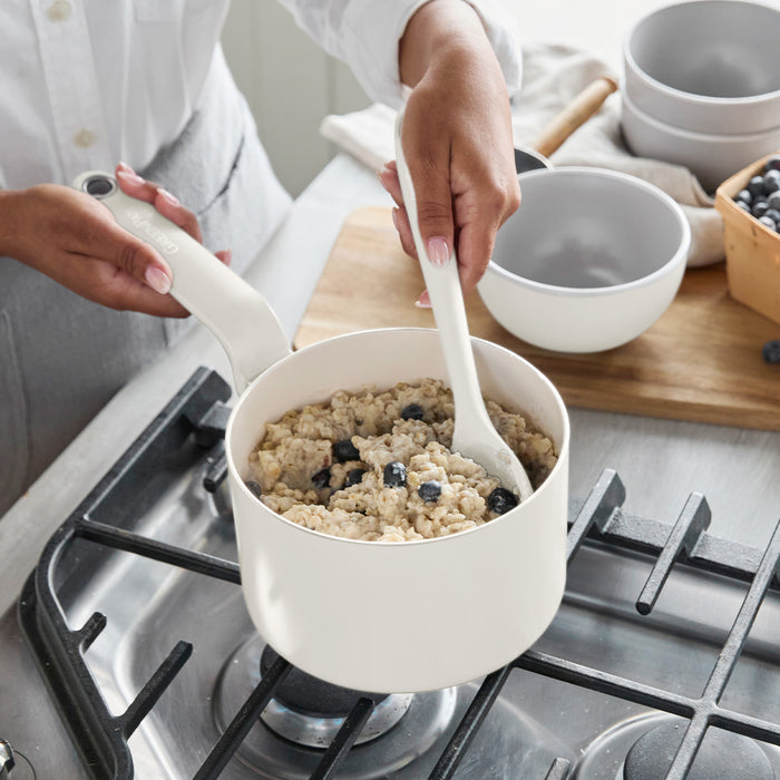 Blueberry oatmeal being stirred in a ceramic pot on the stove, with bowls and fresh blueberries nearby.