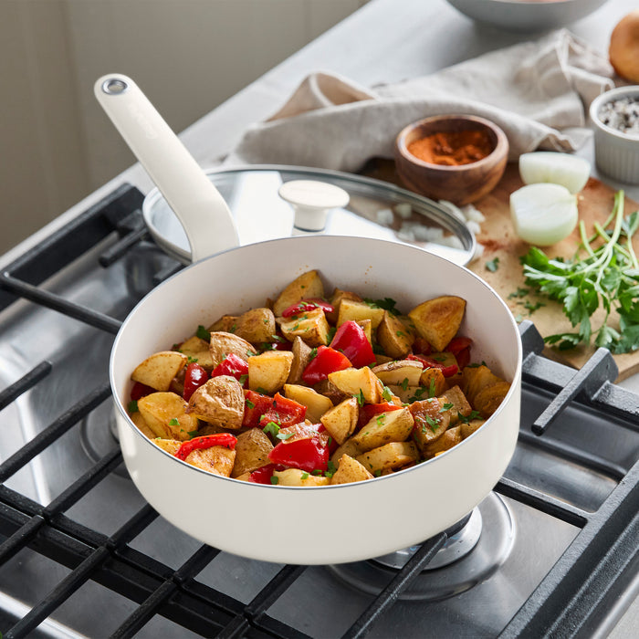 Cream ceramic nonstick 10-piece cookware set on a stove, some pieces filled with roasted potatoes, peppers, and fresh herbs.