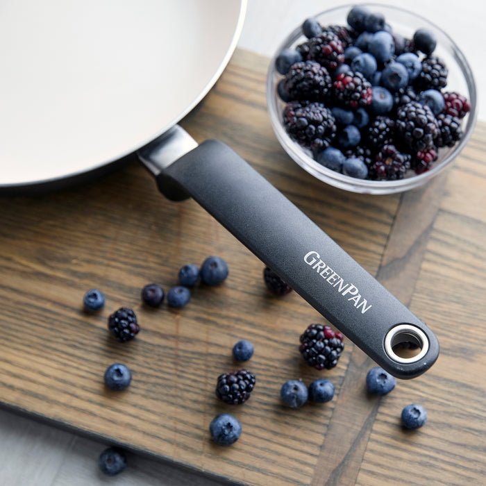 Rio 12-piece ceramic nonstick cookware skillet beside a bowl of mixed berries on a wooden surface, with berries scattered nearby.