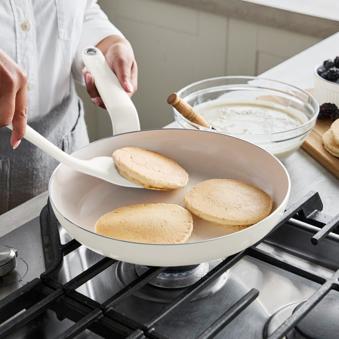 Pancakes being flipped in a ceramic nonstick pan, with pancake batter and fresh berries on the counter nearby.