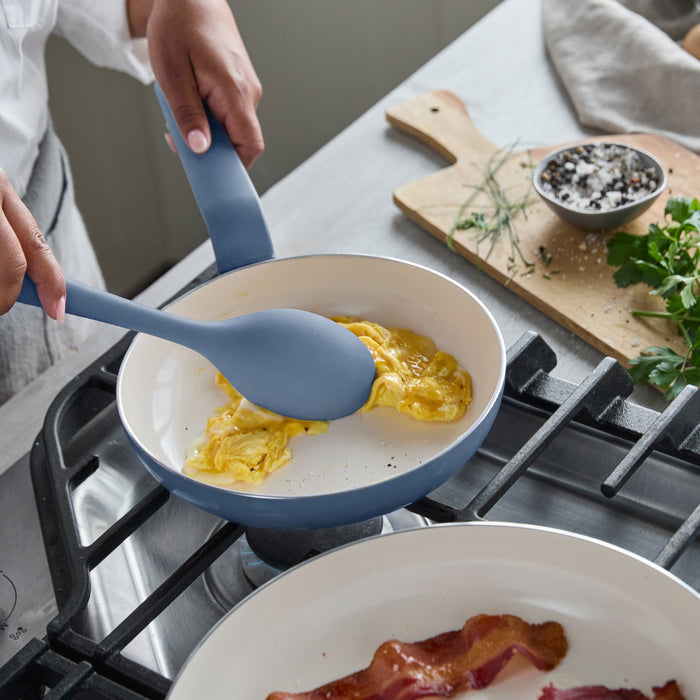 Scrambled eggs cooking in a blue ceramic nonstick pan, with cooked bacon strips and fresh herbs beside the pan.