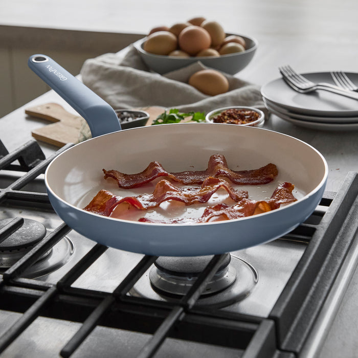 Blue ceramic nonstick cookware set with bacon, eggs, plates, and seasonings arranged on a stovetop.