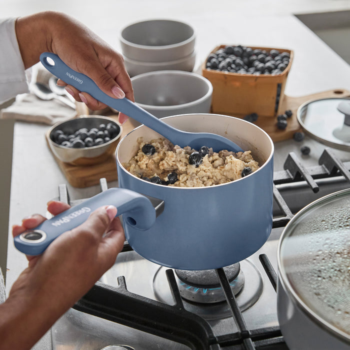 Oatmeal with blueberries being stirred in a blue ceramic pot; matching bowls and fresh berries are on the counter nearby.
