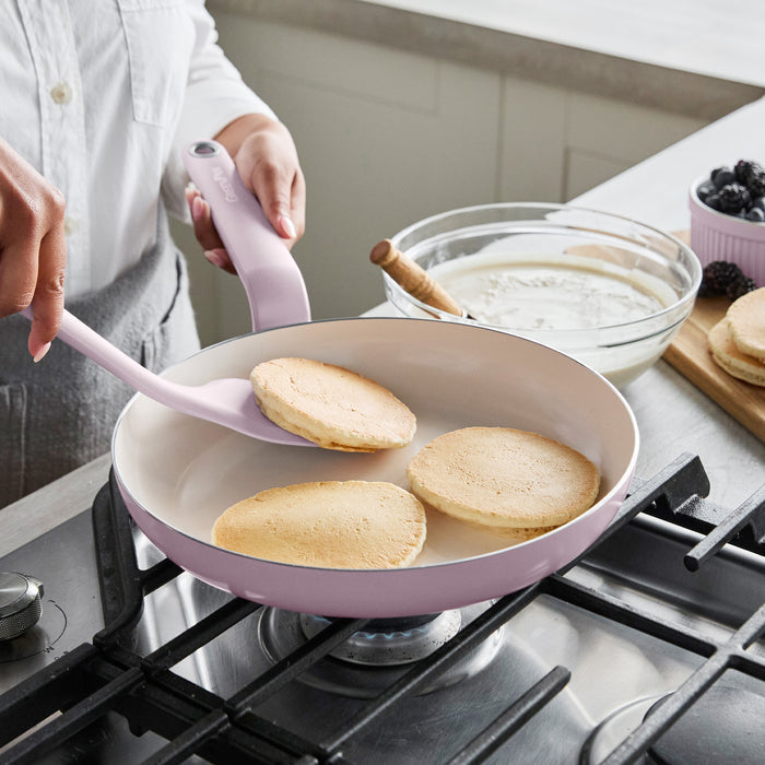 Pink ceramic nonstick pan flipping pancakes, with bowl of batter and scattered berries beside the pan.