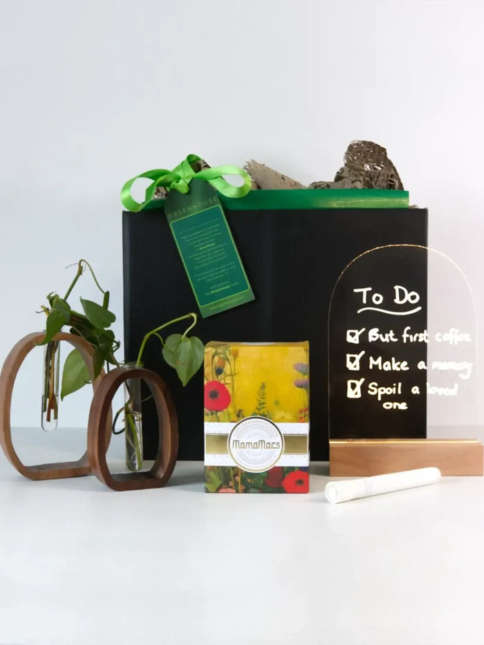 Roots & Rituals Hydroponic Plant Gift Set featuring a black box with a green ribbon. In front, a Pothos plant in two twin wooden holders with glass tubes, a box of Mamamac botanical shortbread biscuits, and a light-up sign board with "To Do: But first coffee, Make a memory, Spoil a loved one" written on it. A chalk paint fine liner rests in front of the board.