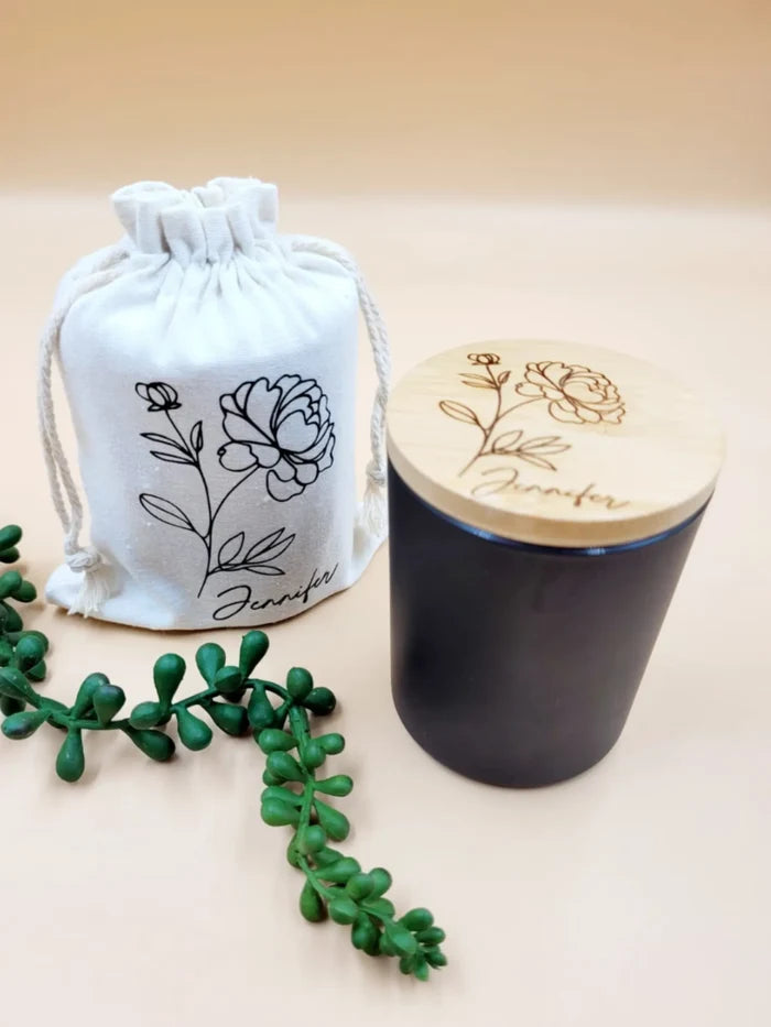 Black candle with wooden lid and white drawstring bag featuring floral designs on a beige background.