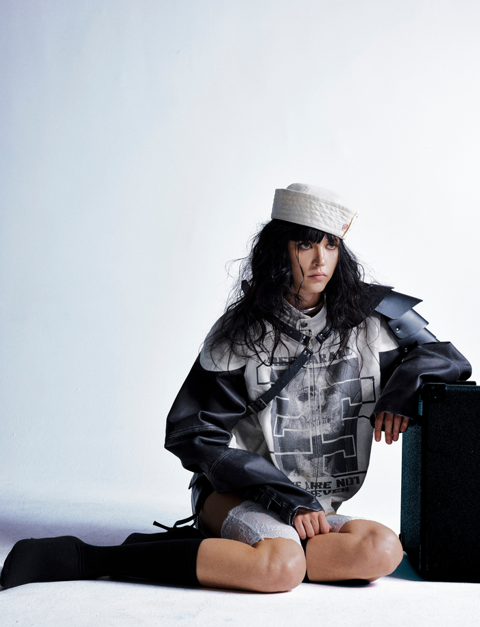 Model sitting with cream and black jacket draped over one arm and a black case beside her