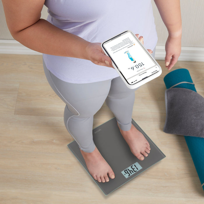 Person standing on a smart scale while holding a smartphone displaying the weight tracking app