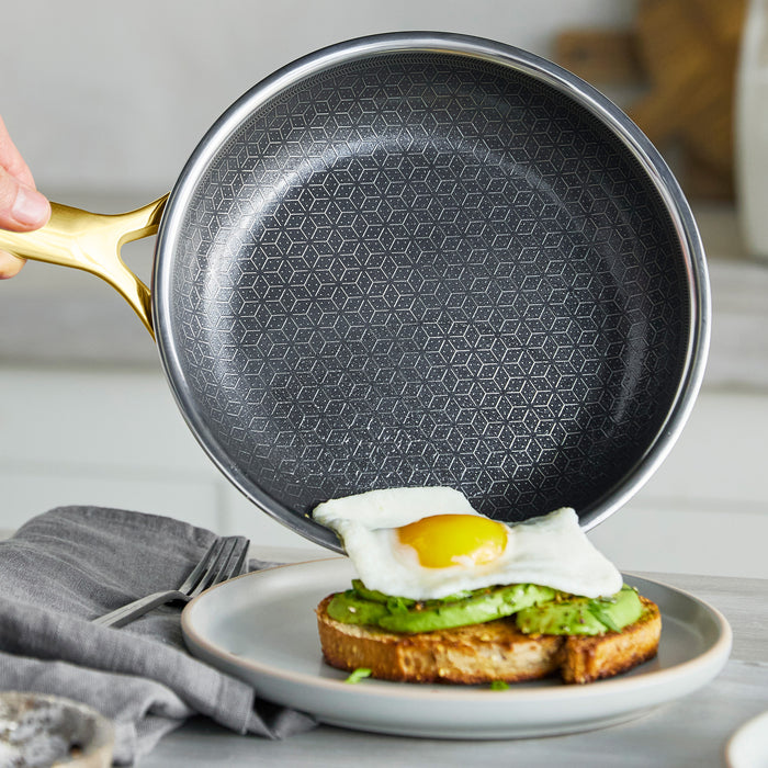Gold ceramic nonstick frypan held above avocado toast topped with a sunny-side-up egg.