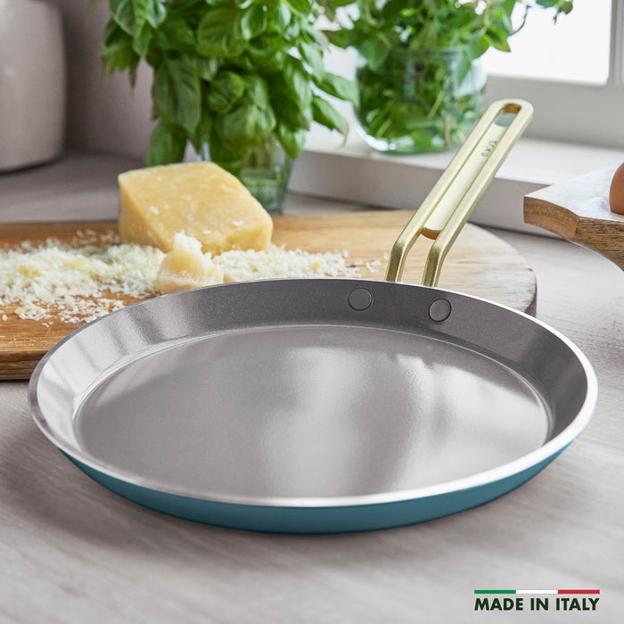 Teal crepe pan on a kitchen counter with cheese and herbs in the background