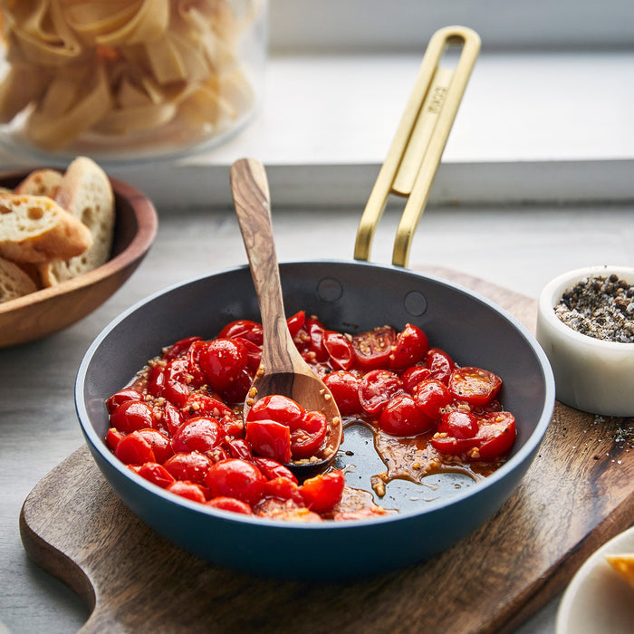 Three-piece ceramic nonstick mini cookware set sautéing tomatoes, with sliced bread, a pepper grinder, and wooden spoon nearby.