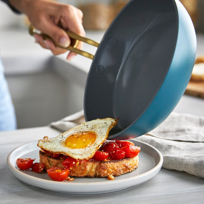 A hand slides a fried egg from a white ceramic nonstick mini pan onto toast topped with cherry tomatoes.