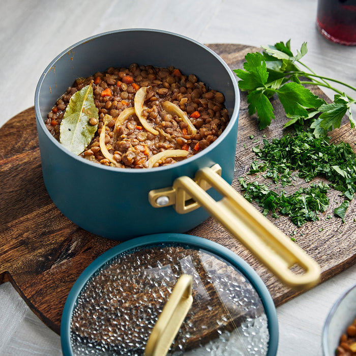 Ceramic nonstick mini cookware set in Venetian Teal with dry lentils, onions, carrots, and parsley beside the pans.