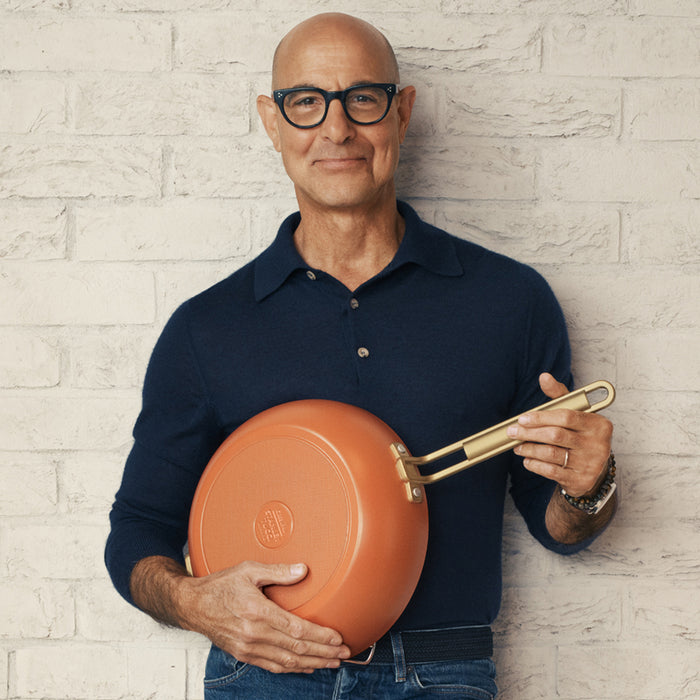 Man in glasses and navy shirt holds a Stanley Tucci 4.5-quart pan with orange accents near a light brick wall.