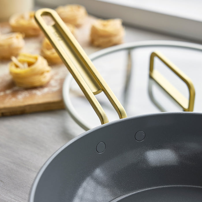 White ceramic nonstick 4.5-quart pan with gold handles and lid, pasta nests placed beside it.