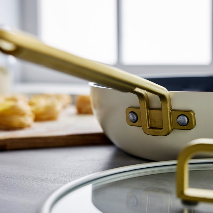 White Stanley Tucci 4.5-quart pan with gold handle and glass lid on a kitchen counter.