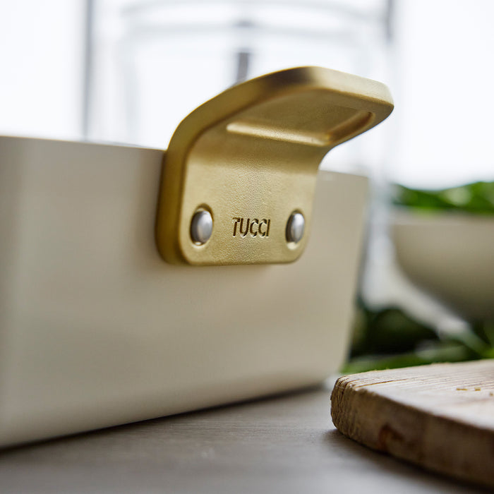 Gold handle on white ceramic lasagna pan with green herbs nearby.